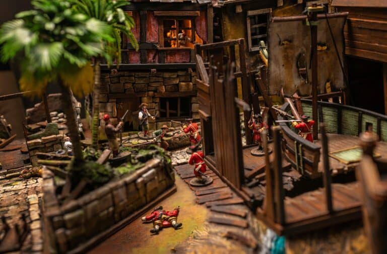 A vibrant tabletop diorama features a detailed pirate scene with miniature figures from Firelock Games. Pirates in red uniforms engage in battle amid rustic buildings reminiscent of Port Royal, ship elements, and lush greenery. A warm lantern glow highlights the rugged, immersive environment.