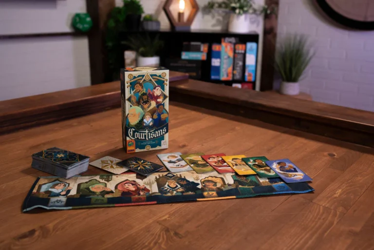 A wooden table holds a colorful card game titled "Courtisans" from Pandasaurus Games, with character cards artfully spread beside the box. The background showcases a shelf filled with more board games, crafting a cozy atmosphere perfect for playful gatherings.