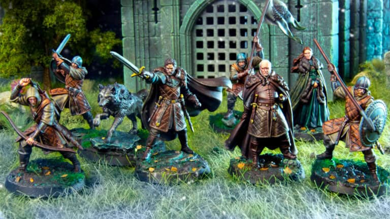 A group of intricately painted medieval warrior miniatures, reminiscent of a Game of Thrones battle, is arranged on a grassy battlefield. Characters wield swords, shields, and spears, with detailed armor and cloaks. A building with an arched gate and bars looms in the background.