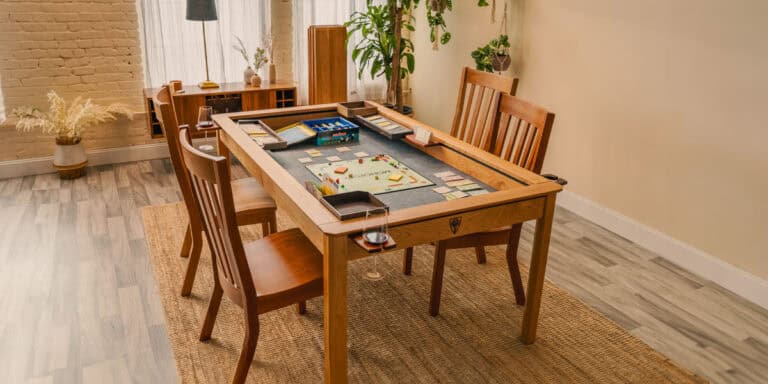 In this cozy room, a modular gaming table is set up for board games. Four wooden chairs surround it, with cards and dice ready for action. Wine glasses sit atop the customizable setup, while a soft rug lies beneath. Sunlight filters through sheer curtains, casting a warm glow over this Kickstarter gem.