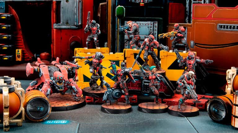 A group of painted miniatures from the March Releases in dynamic poses are set on a tabletop battlefield. They are futuristic soldiers in red armor with various weapons, positioned among industrial-themed terrain with crates and barrels, enhancing your Infinity Roster.