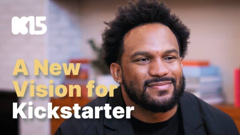 Smiling individual with curly hair and beard in a room, wearing a black jacket. Text on the image reads "A New Vision for Kickstarter" alongside a "K15" logo. This visionary moment highlights the platform's dedication to funding and empowering creators. Background shows books and a plant.