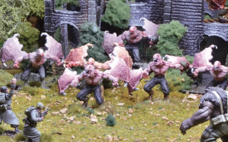 Amid the grassy, rocky terrain, a band of miniature winged creatures with muscular bodies and pinkish wings advances toward a ruined stone building. Below, two figures confront them in a tense standoff reminiscent of an epic Konflikt '47 scenario designed by Andy Chambers for Warlord Games.