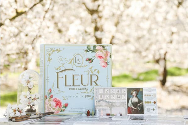 A board game titled "Fleur" is elegantly displayed on a table, capturing the French flair against a backdrop of blooming trees. The game box boasts floral designs, with pieces, cards, and a small figure set up before it. A glass dome filled with flowers adds charm, hinting at an upcoming Kickstarter campaign.