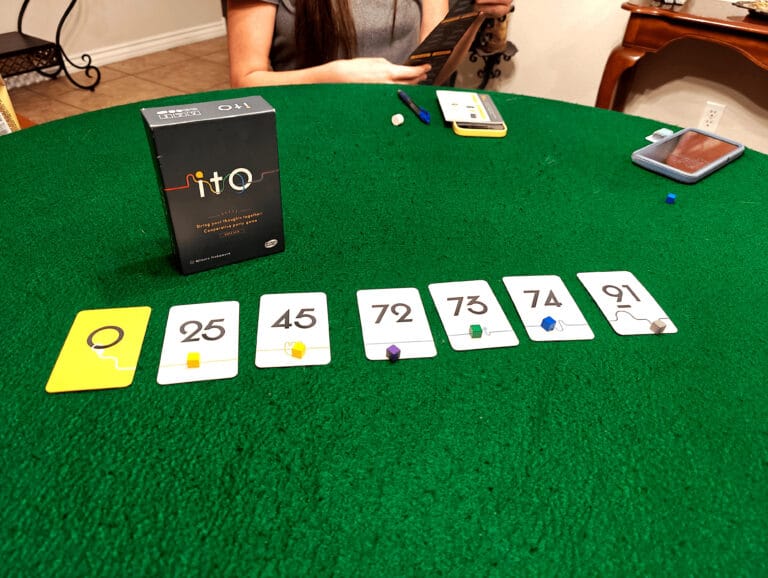 A green table displays a card game set up, featuring a box labeled "The Mind" by Arclight Games and several numbered cards (25, 45, 72, 73, 74, 91) each topped with colored markers. A person holding cards is seated at the table.