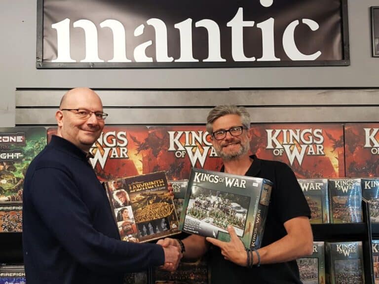 Two people shake hands in front of shelves filled with "Kings of War" game boxes. One holds a "Labyrinth" game and the other a "Kings of War" box, symbolizing a strategic partnership. A "mantic games" sign is visible above them, indicating collaboration with River Horse.