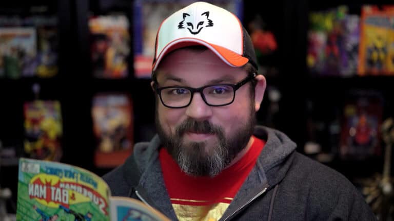 A man with a beard and glasses, wearing a red and white cap with a cat logo, smiles while holding a colorful comic book. Seated in front of a blurred background filled with comic book covers, he resembles SmiteWorks Chief Development Officer Adam Bradford.