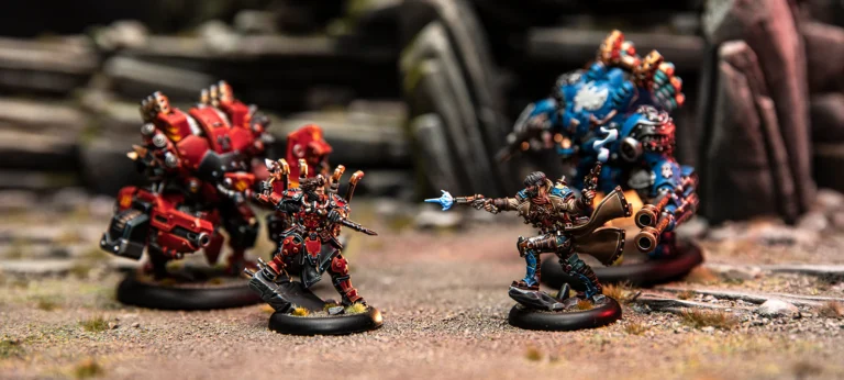 Miniature figures depicting a dynamic battle scene on a detailed terrain. Two vividly painted humanoid robots from Warmachine, one red and one blue, face off with two smaller, armored characters poised to attack. The background reveals stone ruins reminiscent of the Iron Kingdoms.
