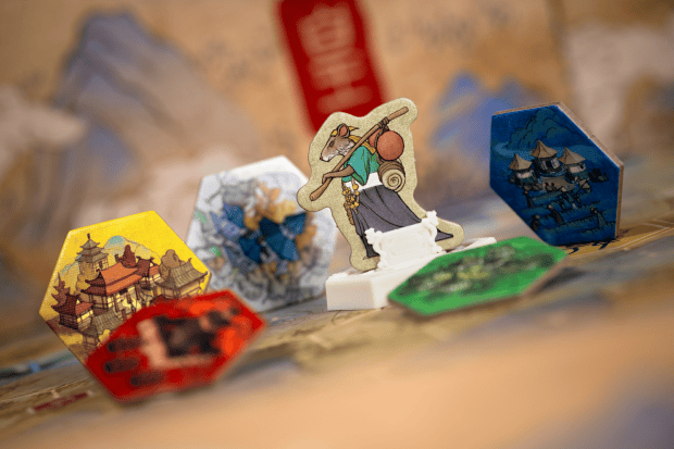 The board game pieces rest on a vibrant game board, featuring colorful hexagonal tiles with intricate designs. A central figurine of a character with a staff and scroll stands out in the foreground. Designed by Reiner Knizia, the game highlights his trademark strategic depth and elegance.
