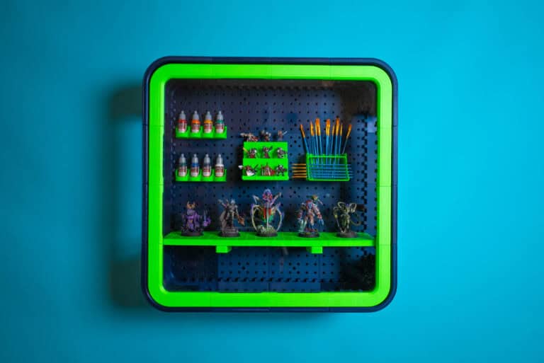 A wall-mounted JudoGrid display case with a black pegboard showcases small paint bottles, miniature figures, and paintbrushes on bright green shelves against a blue wall. The innovative modular display system was successfully launched on Kickstarter.