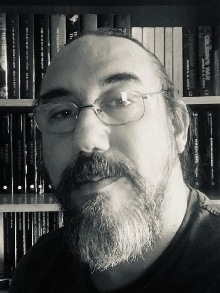 A black and white close-up of a bearded person with glasses, possibly an enthusiast of Gav Thorpe's works, standing in front of a bookshelf filled with books that hint at the tactical depth found in Warlord Games.