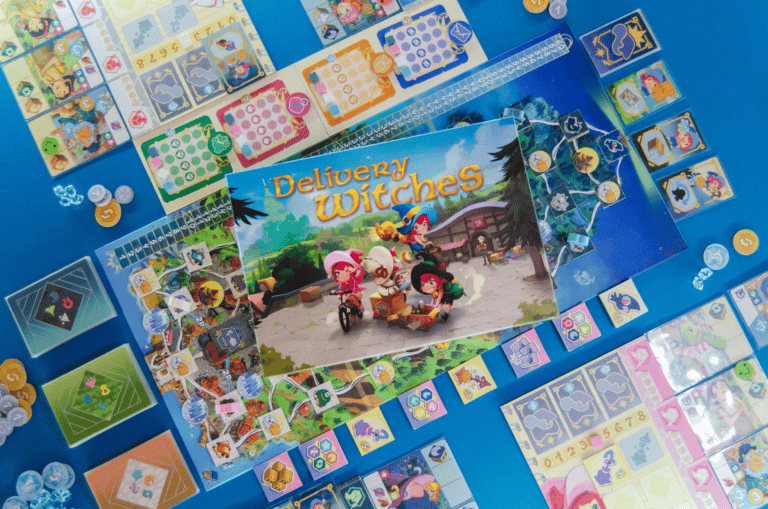 A colorful board game setup featuring the Kickstarter sensation "Delivery Witches" with cards, tokens, and game boards arranged on a blue surface. The cover art showcases animated witches in a whimsical setting, mastering their witch apprenticeship while casting enchanting spells.