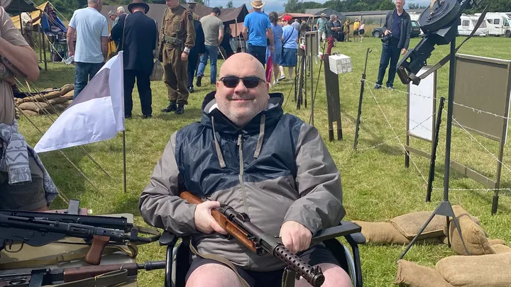 At an outdoor historical reenactment event, Paul Sawyer, a veteran in a wheelchair, is holding a rifle. The grassy field is alive with various people and vintage military equipment in the background, capturing the spirit of wargaming and history alike.