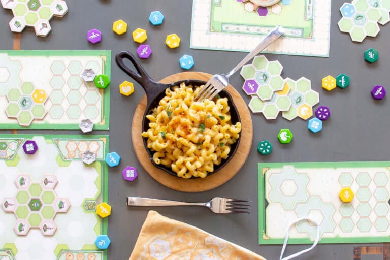 A skillet of macaroni and cheese sits on a table at Mox Boarding House, surrounded by colorful board game pieces and tiles. A fork is in the dish, with another fork and a napkin nearby. The scene exudes a playful yet cozy atmosphere, merging gastronomy with the joy of tabletop games.