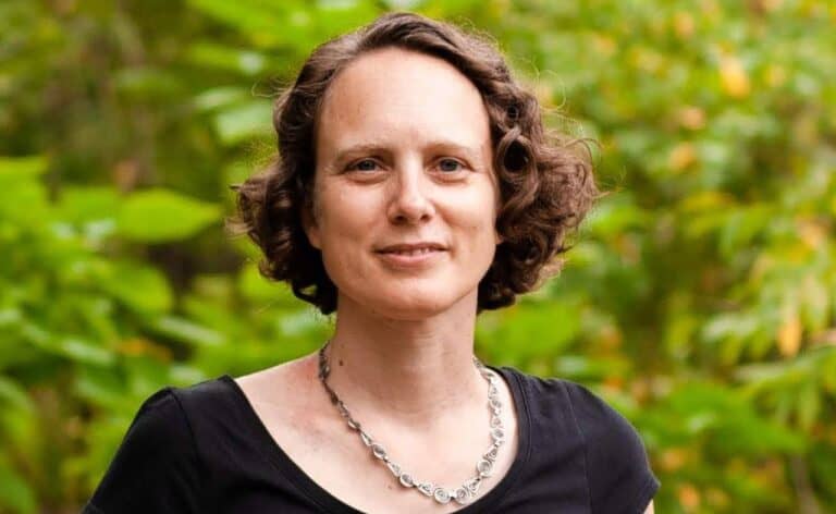 A person with short curly hair, wearing a black shirt and a necklace, stands outdoors in front of green foliage, smiling slightly, reminiscent of Elizabeth Hargrave's relaxed demeanor at PAX Unplugged 2024.
