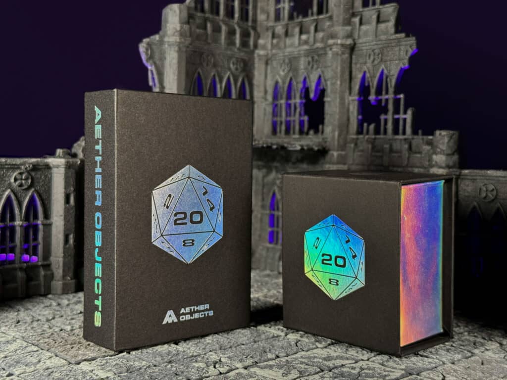 Two black boxes, adorned with colorful holographic Cathedral Dice designs, are set against a gothic-style building backdrop. The larger box reads "Aether Objects," enhancing the dark, mysterious ambiance with subtle purple lighting. Perfect for enthusiasts of Gothic-Inspired RPG Dice.
